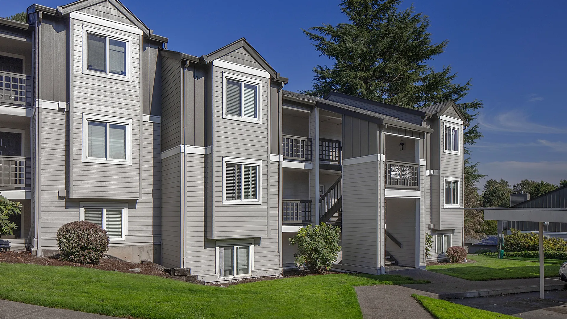 Charming modern apartment exterior with crisp gray siding, lush green landscaping, and picturesque balconies under a clear blue sky.