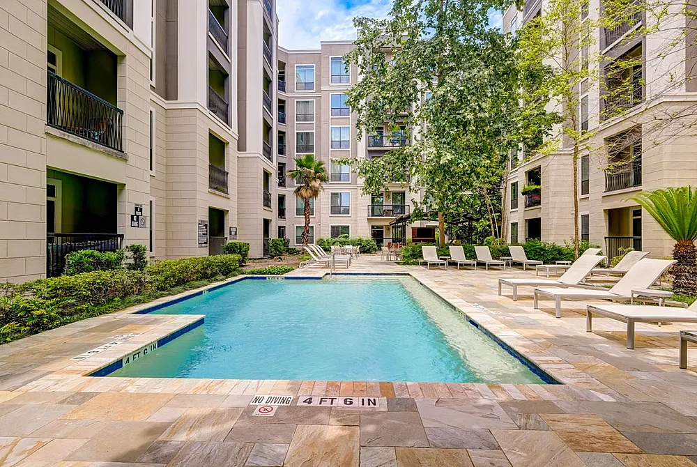 Luxurious apartment complex courtyard featuring a sparkling swimming pool surrounded by lush greenery and elegant lounge chairs for relaxation.