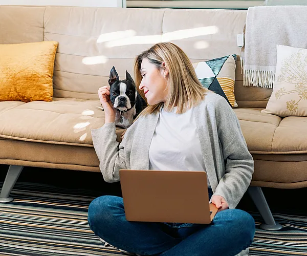 Cozy living room with a modern beige sofa, colorful cushions, and a joyful atmosphere featuring a woman using a laptop and a playful small dog.