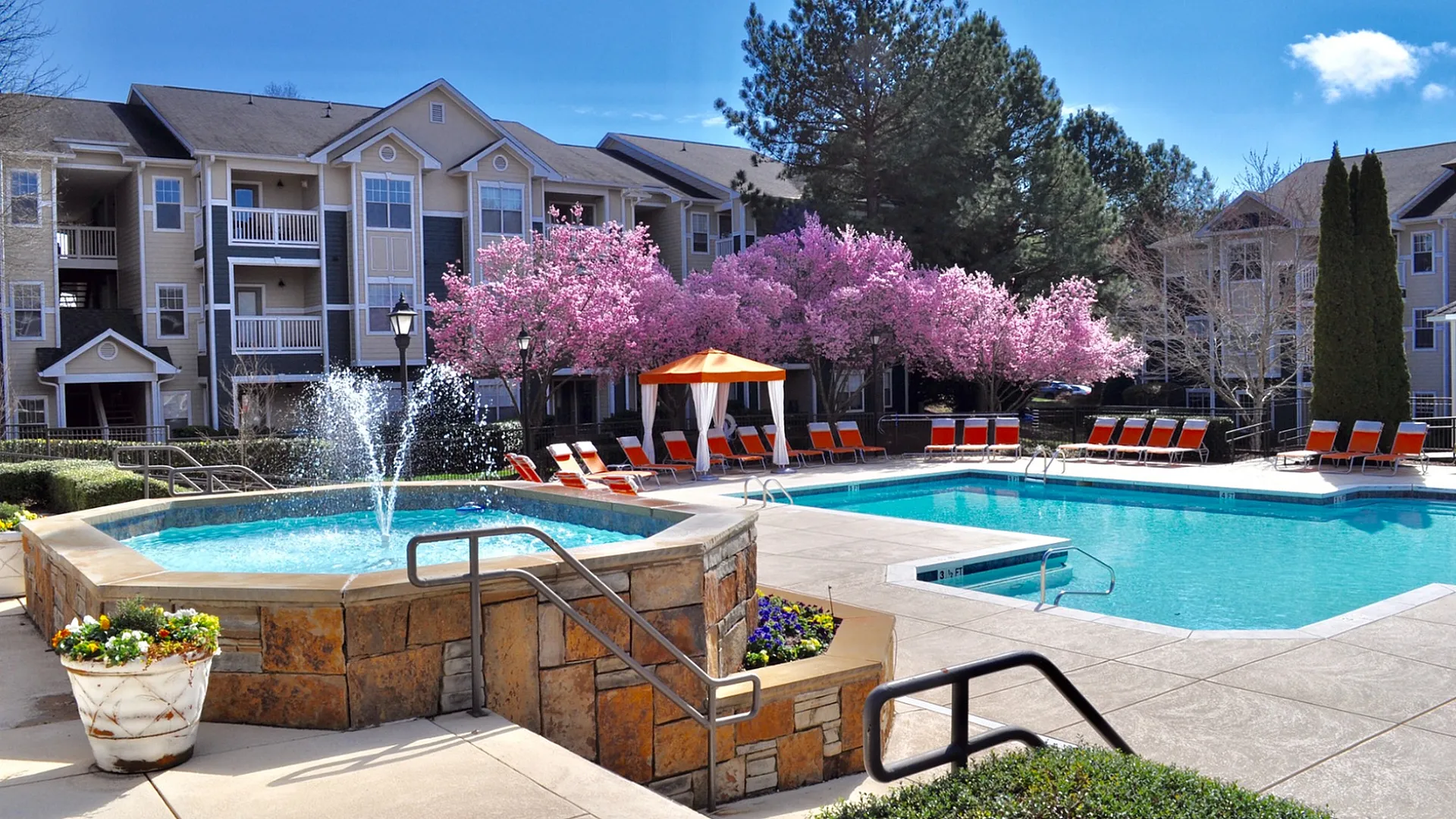 Luxurious apartment complex with a stunning pool area, surrounded by vibrant pink blossom trees and stylish lounge chairs, offering a serene and refreshing atmosphere.