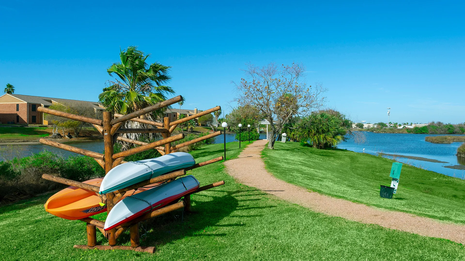 Scenic outdoor area with a vibrant lawn, palm trees, and kayaks overlooking a serene lake beside modern apartments.