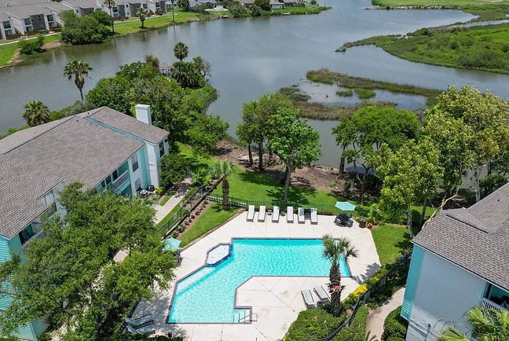 Aerial view of a serene apartment complex featuring a sparkling pool, surrounded by lush greenery and overlooking a tranquil lake.