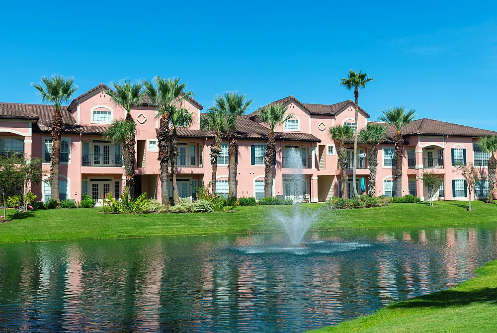 Charming apartment complex with light pink stucco and lush palm trees, overlooking a serene pond with a central fountain.
