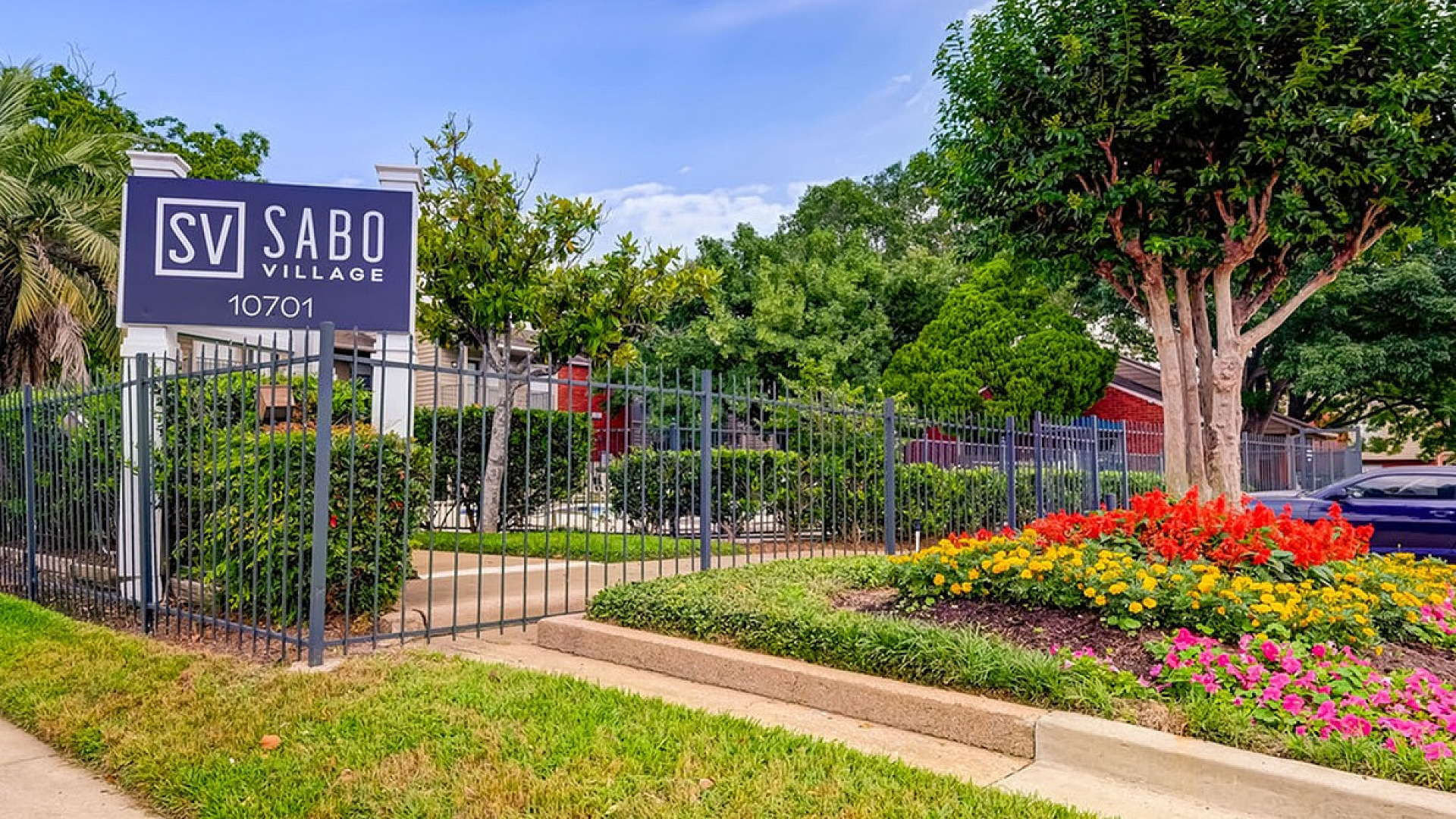 Inviting entrance of Sabo Village with vibrant landscaping, lush greenery, and modern signage under a clear, blue sky.