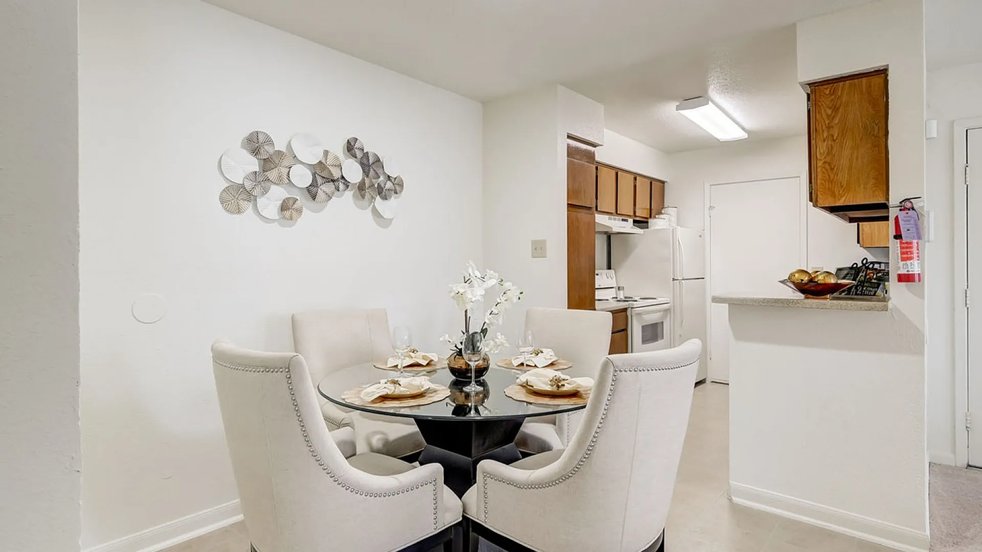 Chic dining area with elegant seating and a glass-top table, adjacent to a cozy kitchen with warm wooden cabinetry.