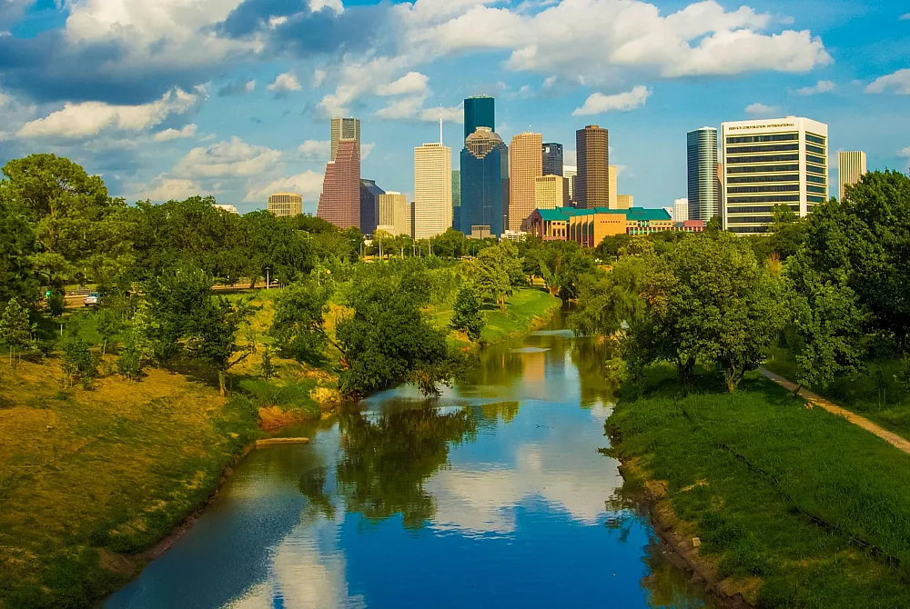 Vibrant cityscape with modern skyscrapers overlooking a lush, green park and serene river under a bright blue sky.