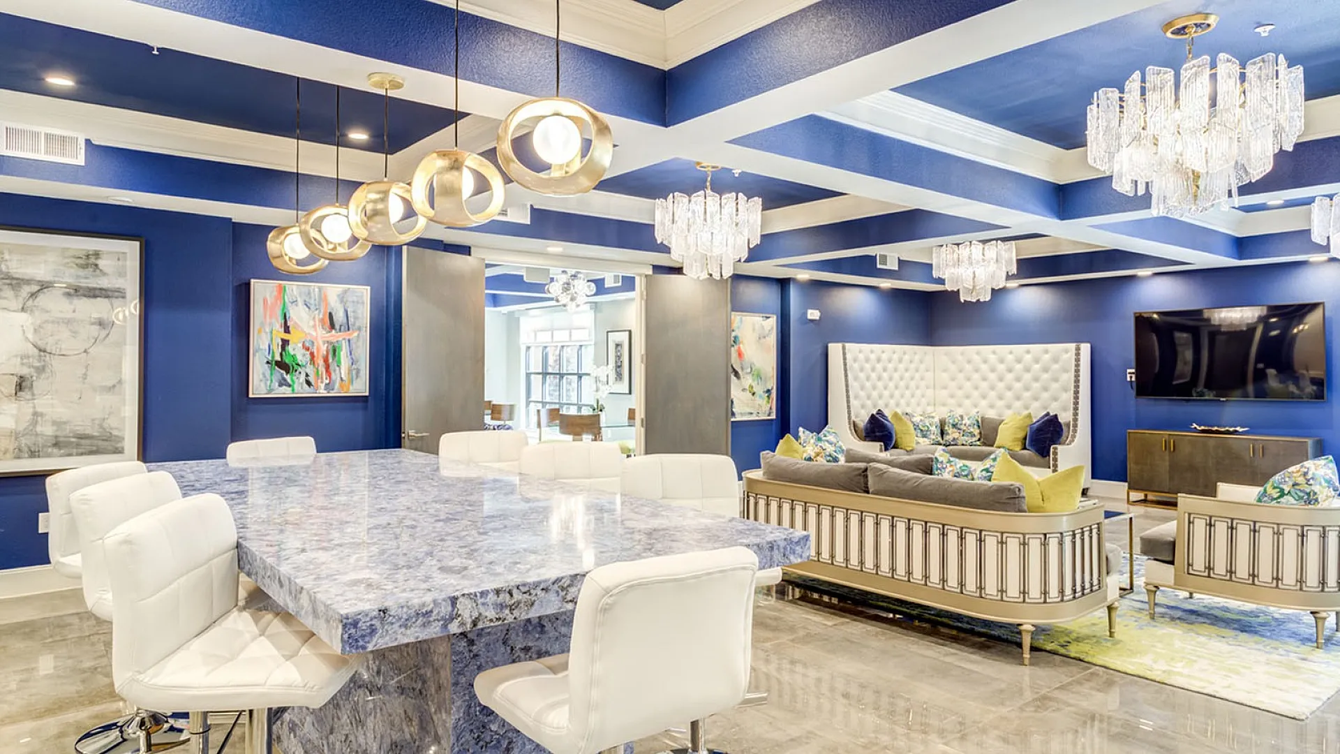 Luxurious lounge area with vibrant blue walls, elegant chandeliers, and modern art, featuring a blue marble-top table and plush seating.