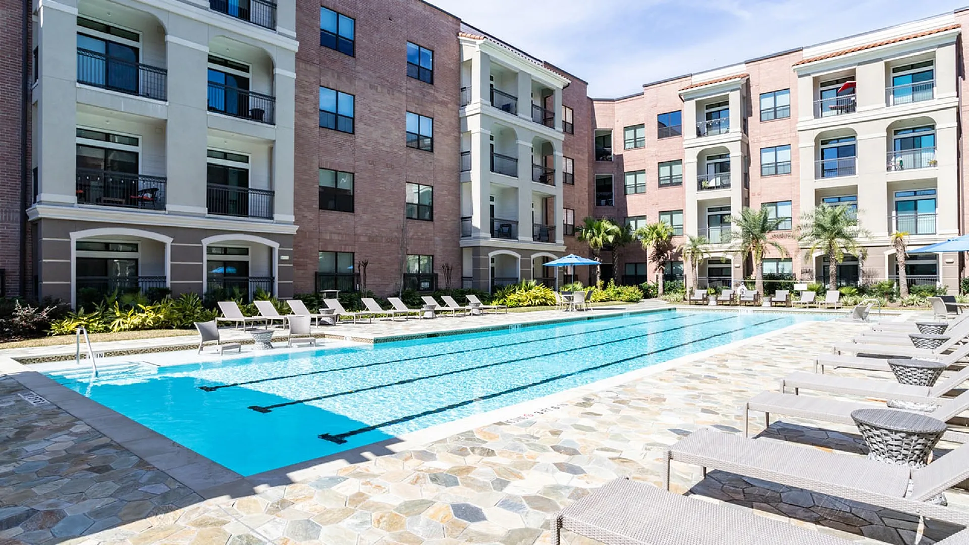 Luxurious apartment complex featuring a sparkling outdoor swimming pool surrounded by elegant lounge chairs, set against a backdrop of modern architecture and lush greenery.