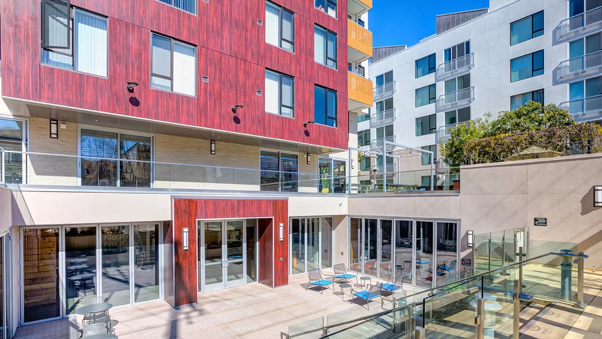 Modern apartment complex with vibrant red wood facade, spacious outdoor seating area, and glass railings.