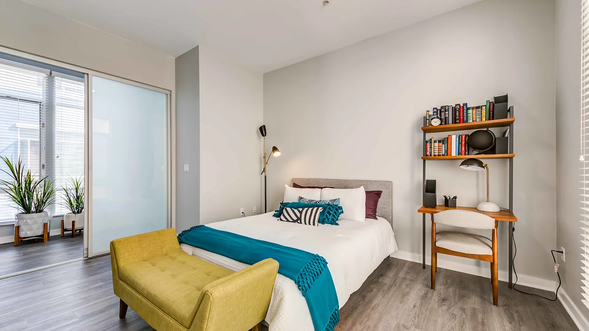 Inviting bedroom with modern decor, featuring a comfortable bed, stylish yellow bench, and a chic bookshelf with desk under soft natural light.