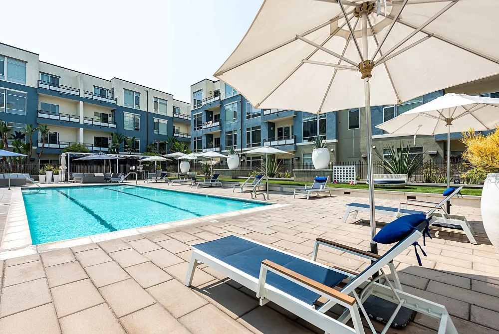 Modern apartment complex with an inviting outdoor swimming pool and comfortable sun loungers, surrounded by lush greenery and elegant umbrellas.