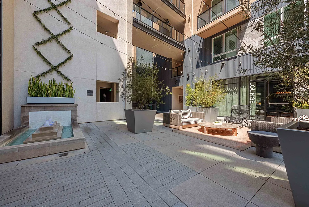 Modern outdoor lounge area with stylish seating, lush greenery, cascading water feature, and elegant string lights.