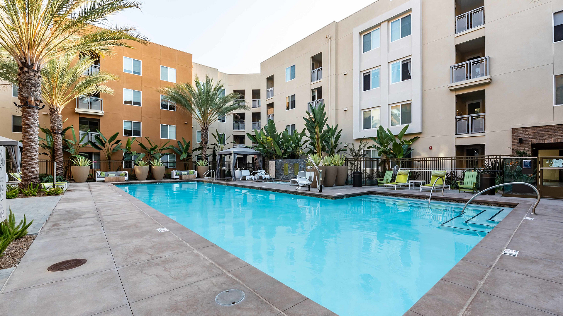 Luxe apartment complex with a sunlit swimming pool surrounded by tropical palm trees and elegant poolside seating.