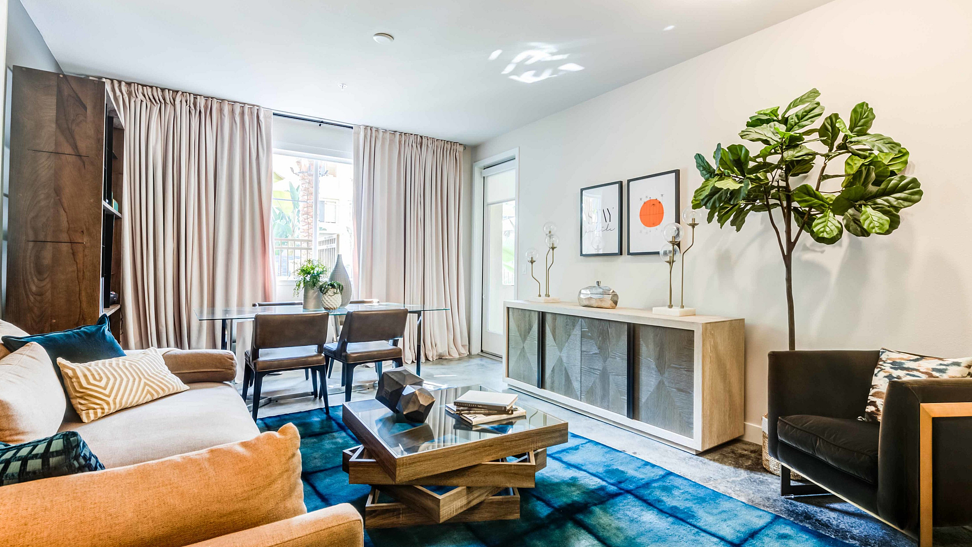 Elegant living room with modern decor, featuring a cozy beige sofa, stylish coffee table, and a vibrant blue rug, complemented by large windows and lush indoor plants.