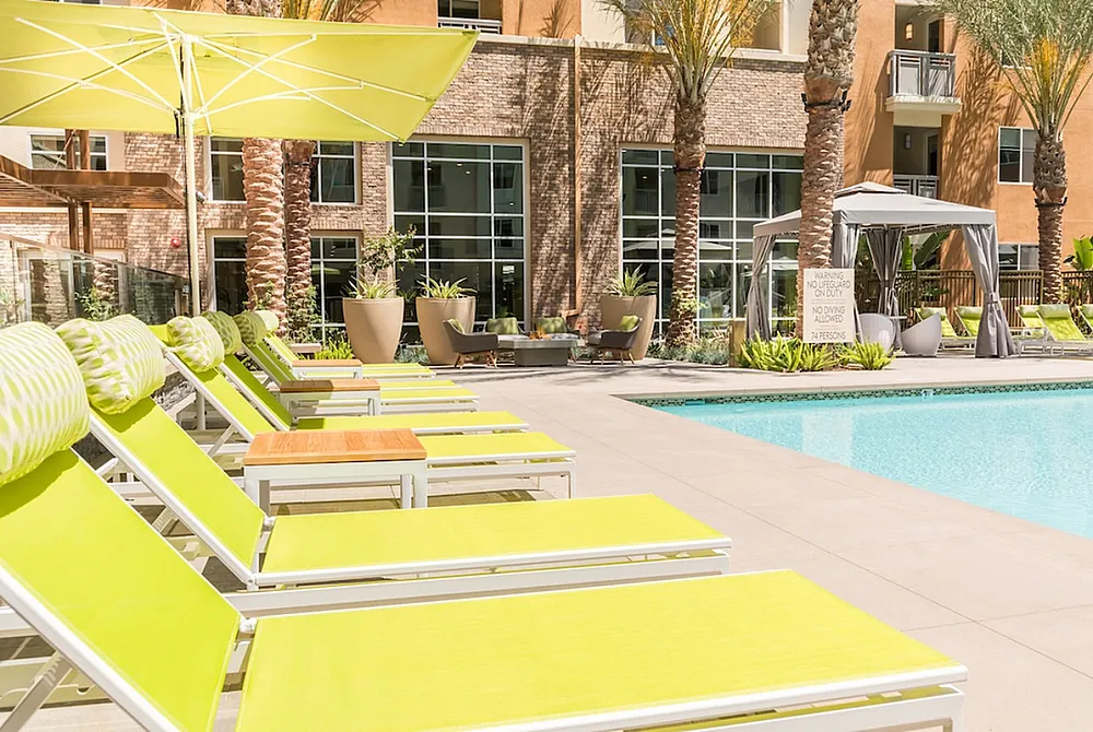 Inviting apartment pool area with vibrant yellow loungers, shaded cabana, and lush palm trees.