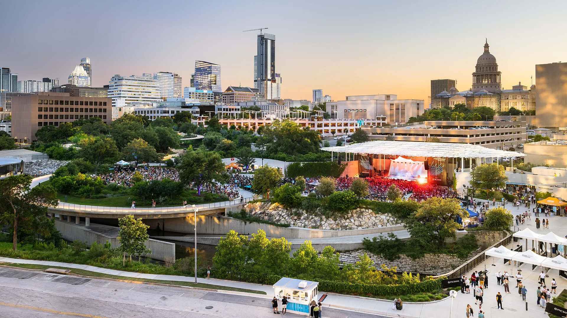 Vibrant cityscape overlooking an open-air concert with a modern downtown skyline, lush greenery, and lively crowd enjoying the event.