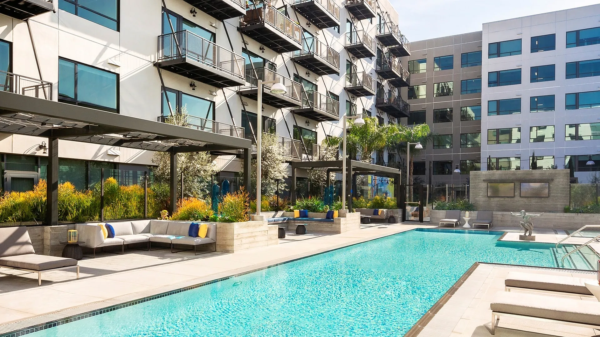 Luxurious modern apartment complex with a sparkling swimming pool, poolside loungers, and stylish outdoor seating surrounded by lush greenery.