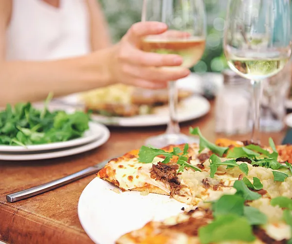 Inviting dining scene with a gourmet pizza topped with fresh greens, accompanied by a glass of white wine and a lush salad, perfect for an alfresco meal.
