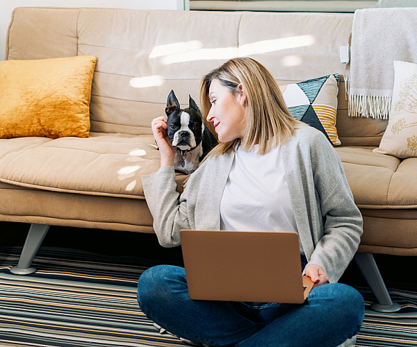 Cozy living room with a woman working on a laptop, enjoying time with her Boston Terrier. Modern design with a stylish beige sofa and decorative pillows.