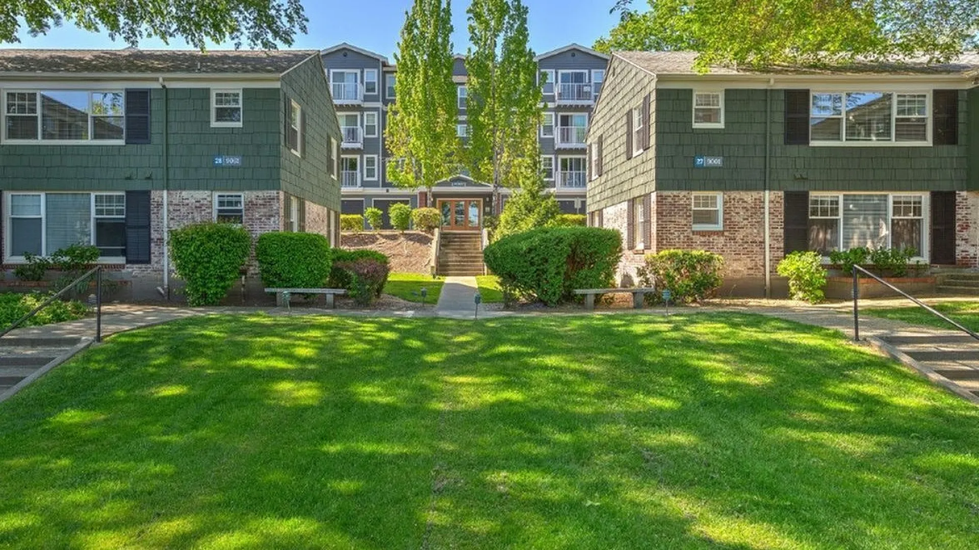 Charming residential complex with lush green lawns, mature trees, and inviting multi-story buildings under a bright blue sky.