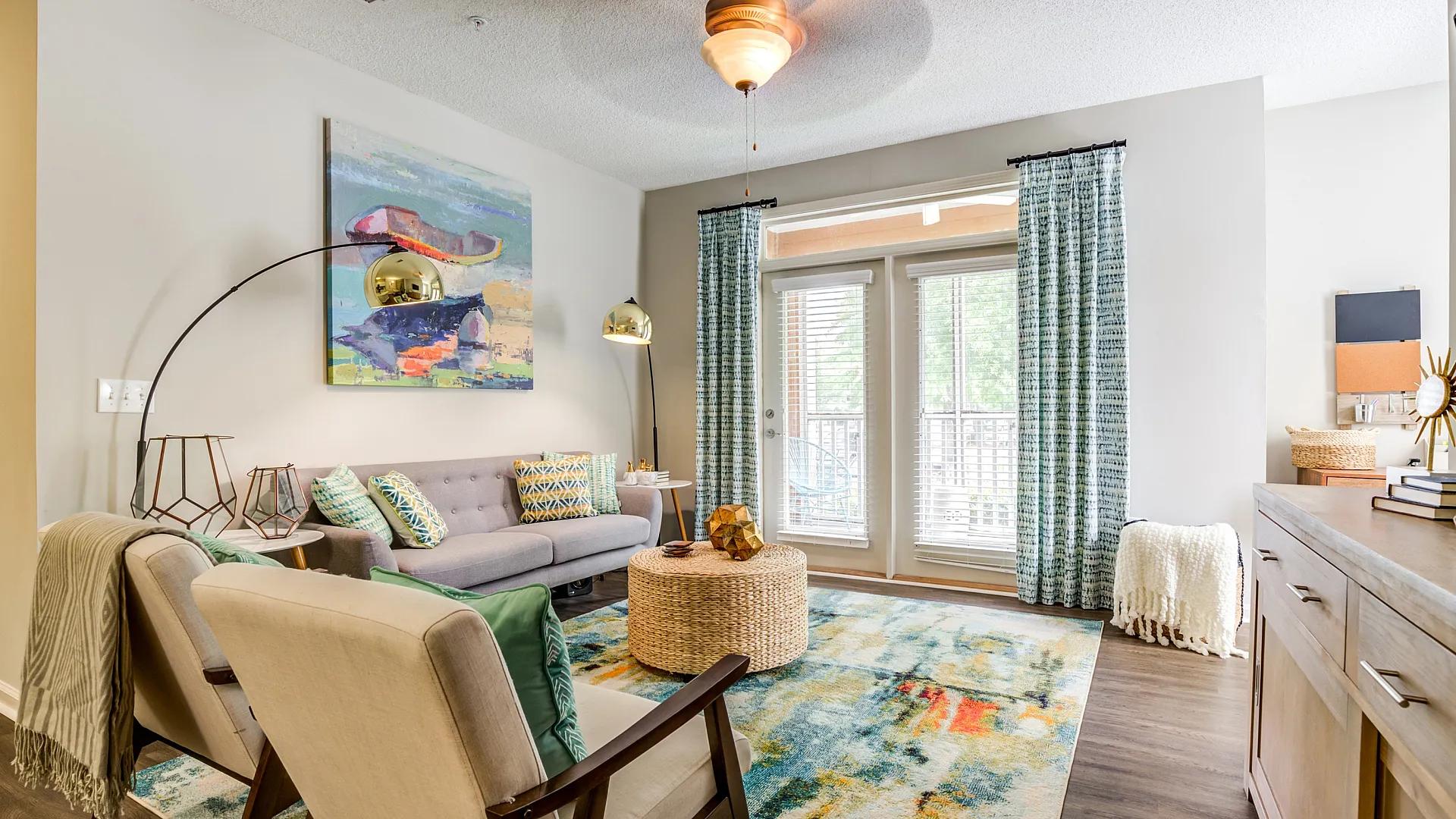 Bright and inviting living room featuring modern decor, a plush gray sofa, vibrant throw pillows, and abstract artwork. Natural light floods through large windows with elegant curtains, highlighting the colorful area rug and stylish furnish
