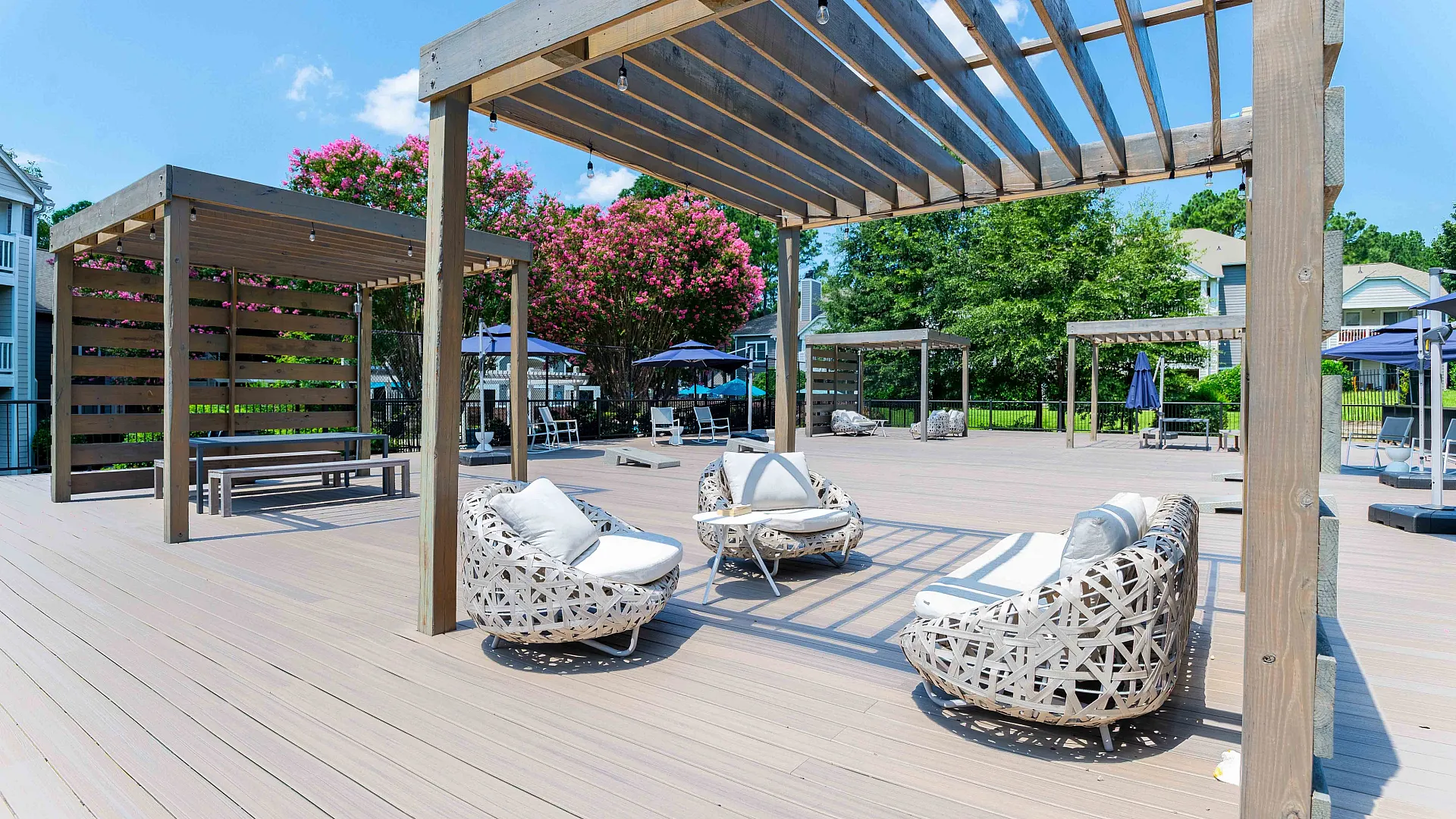 Luxurious outdoor lounge area with stylish wicker seating and wooden pergolas, surrounded by lush greenery and vibrant blooms under a clear blue sky.