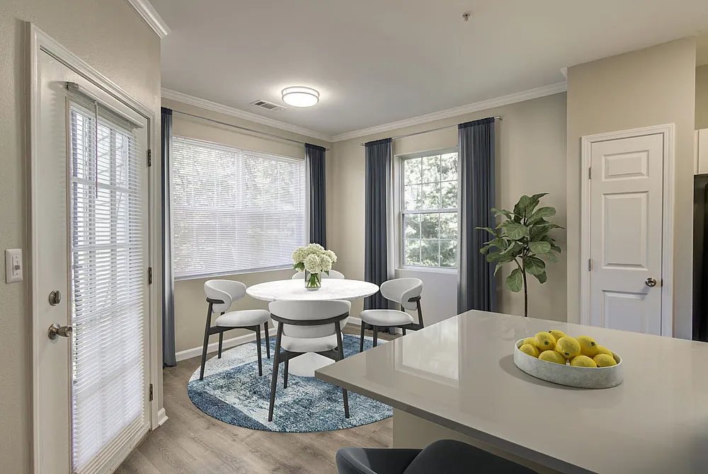 Bright and inviting dining area with a stylish round table, elegant white chairs, and a charming view of greenery through large windows.