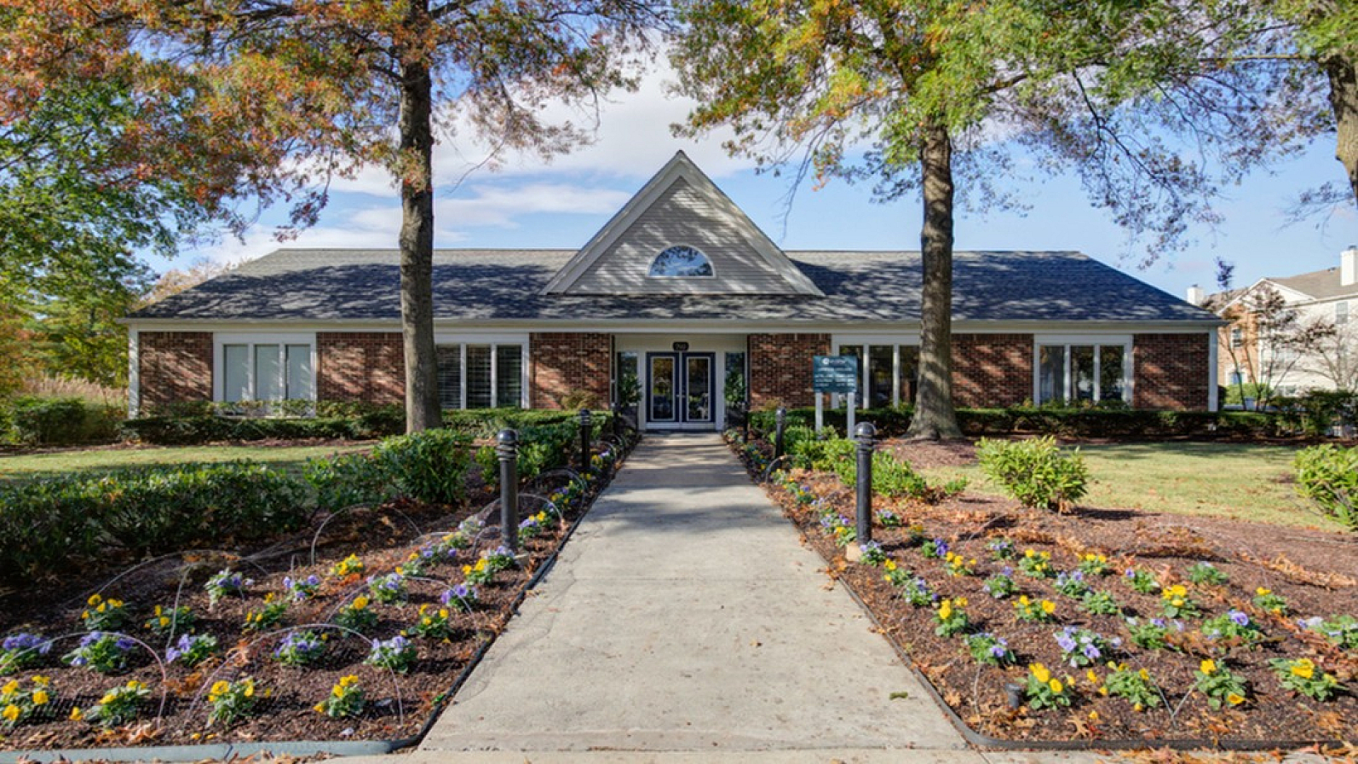 Charming brick building with a picturesque entrance flanked by vibrant flower beds and tall trees, offering a welcoming and serene atmosphere.