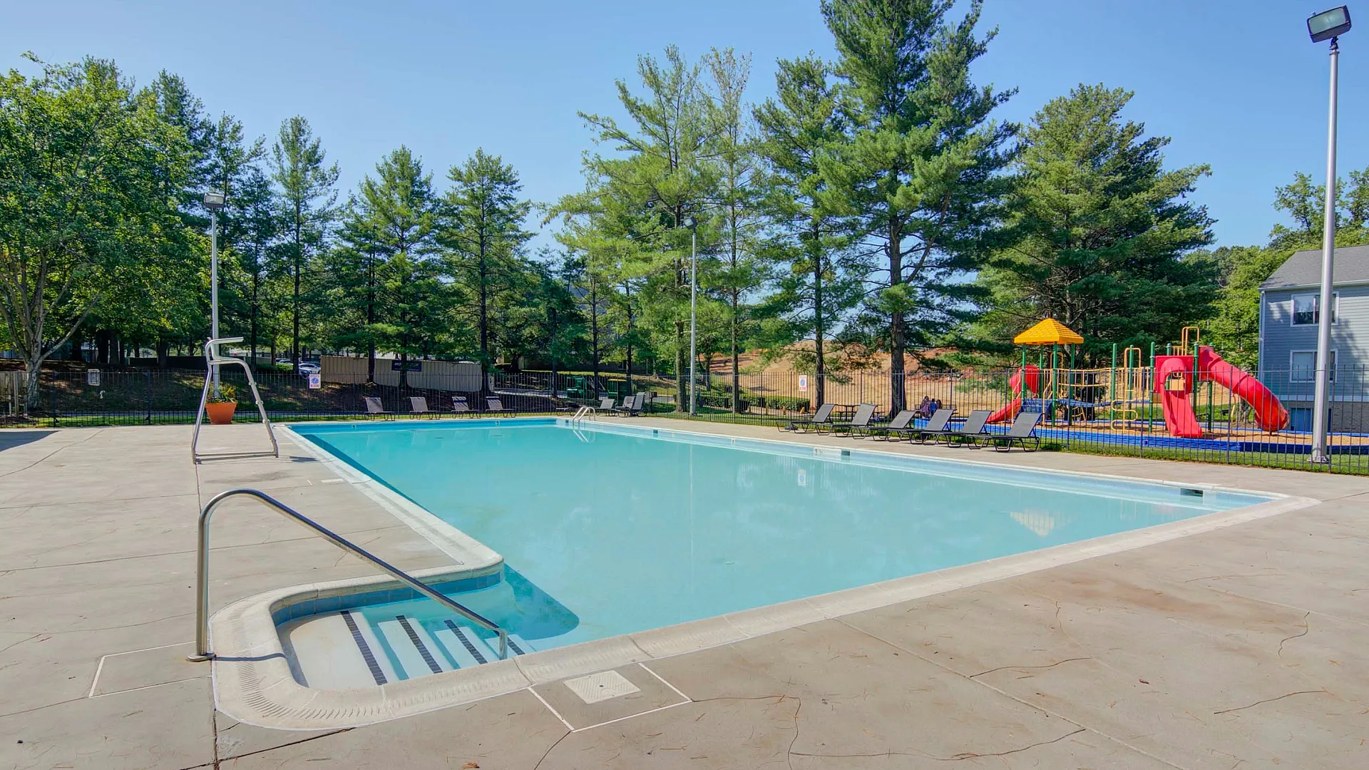 Inviting outdoor pool surrounded by lush trees and comfortable lounge chairs, adjacent to a vibrant playground.