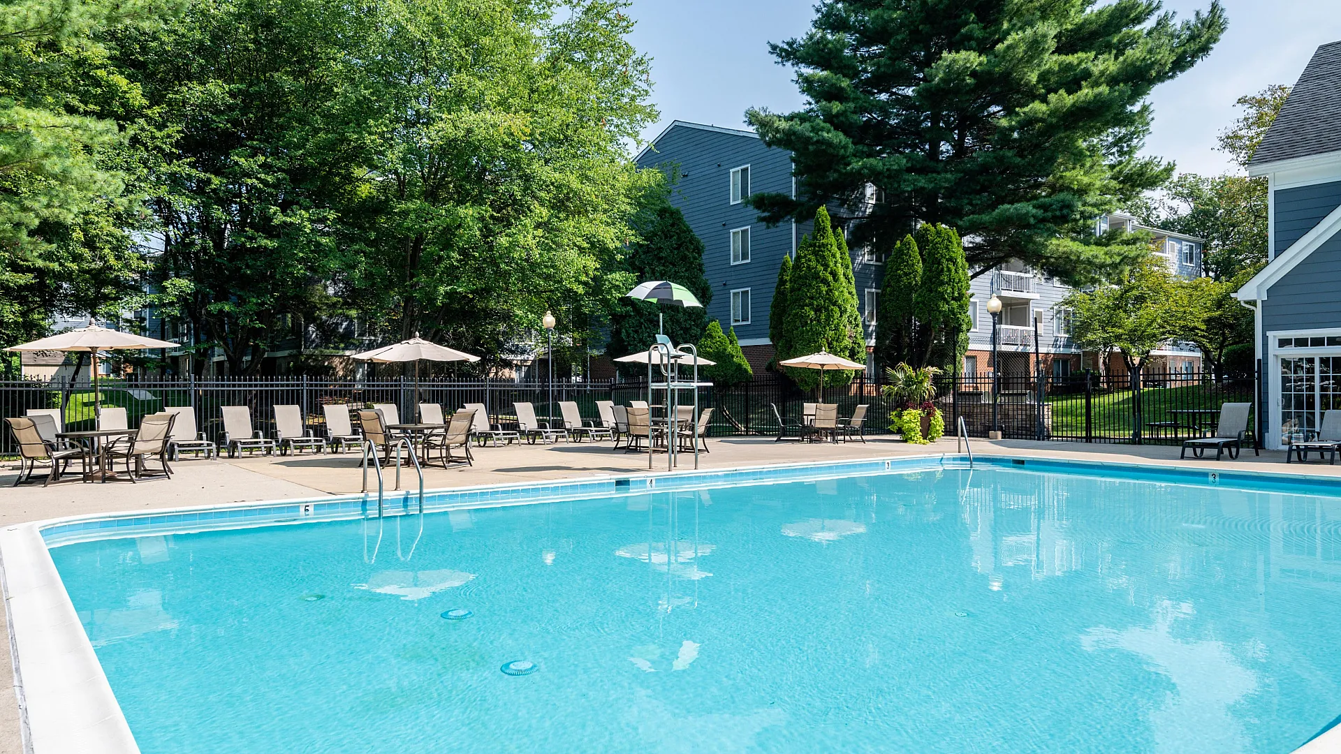 Inviting outdoor pool area with comfortable lounge chairs and umbrellas surrounded by lush greenery in a modern apartment complex.