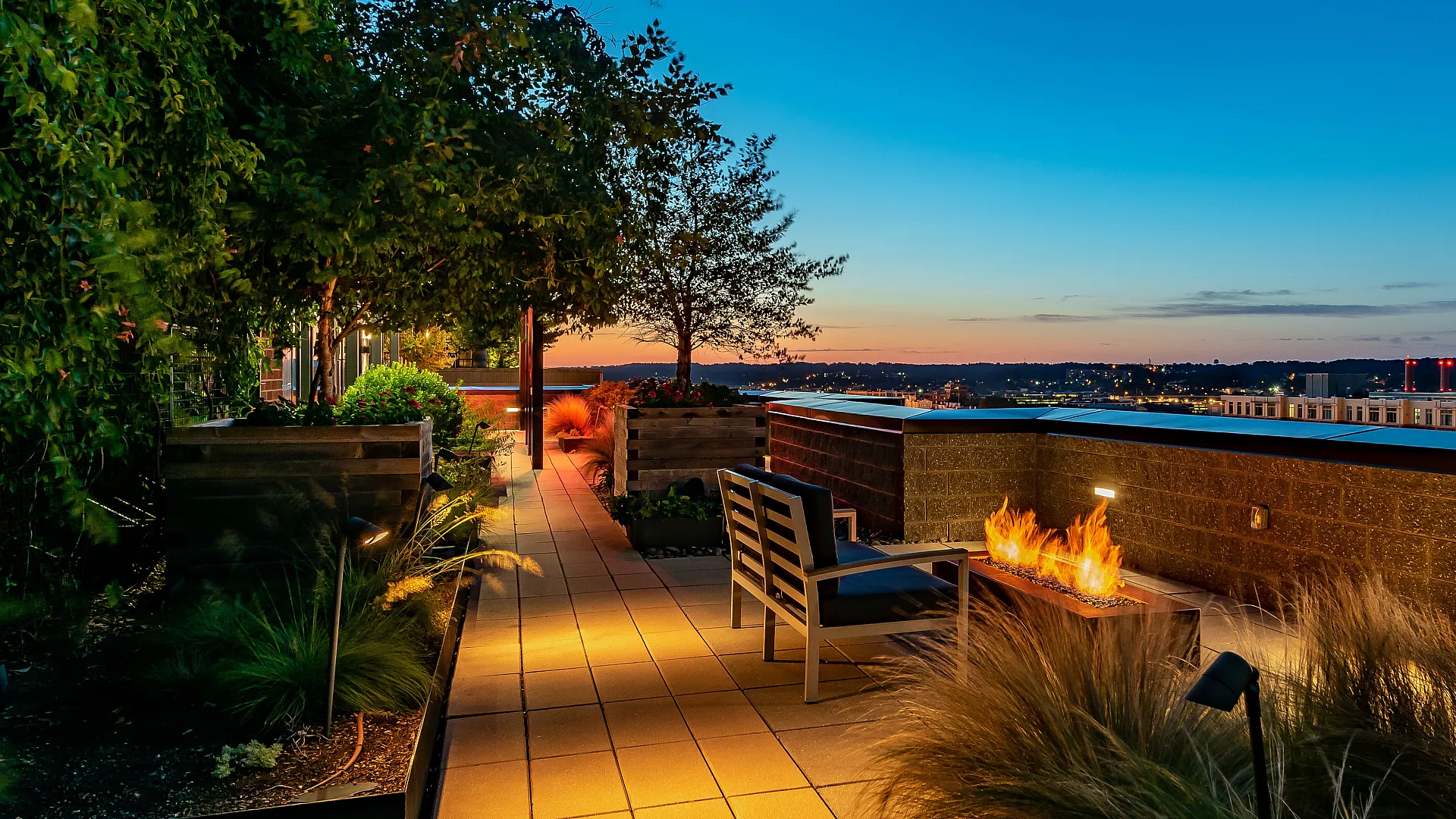 Lavish rooftop terrace with cozy seating, ambient lighting, lush greenery, and modern firepit overlooking scenic city views at sunset.