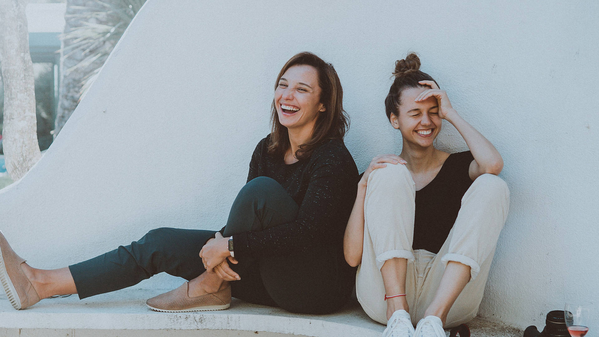 Two women sitting and laughing on a stylish outdoor bench, creating a joyful and relaxed ambiance with a glass of rose wine and camera nearby.