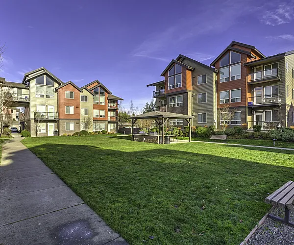 Charming apartment complex with modern architecture, lush green courtyard, and outdoor seating, basking under a clear, bright sky.