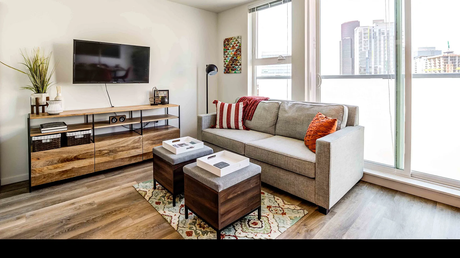 Bright and modern living room featuring a cozy gray sofa with vibrant cushions, large windows with city views, and a stylish media console beneath a mounted flat-screen TV.