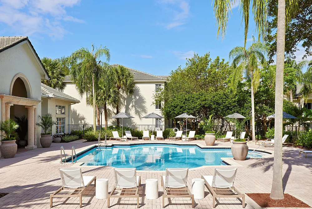 Luxurious apartment complex featuring a sparkling pool surrounded by lush palm trees, elegant loungers, and stylish umbrellas.