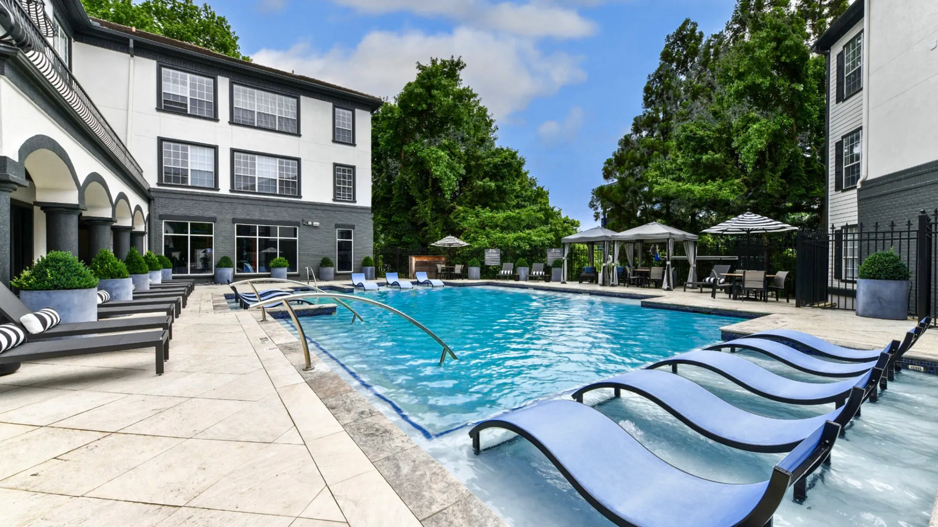 Luxurious apartment complex featuring a sparkling outdoor pool with sun loungers and stylish cabanas set against lush greenery.