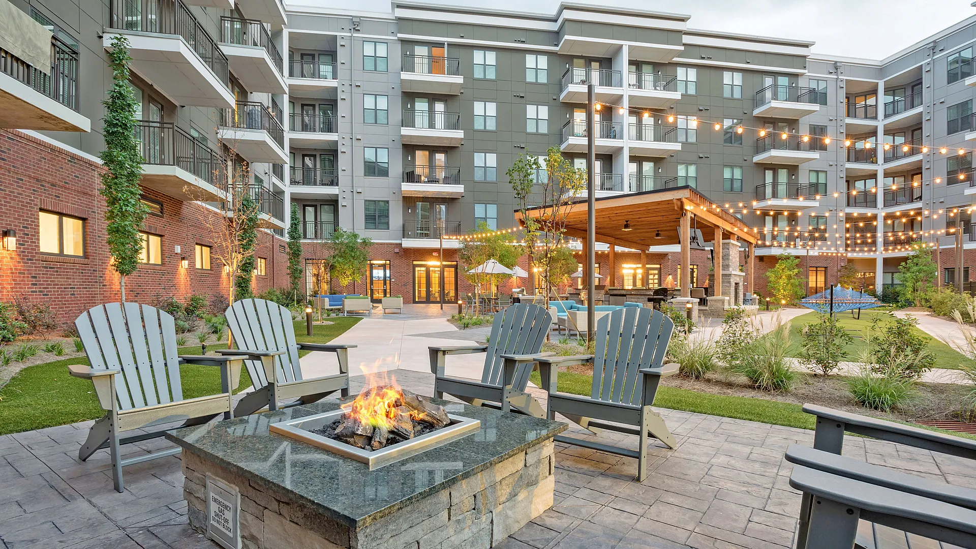 Modern apartment complex courtyard with cozy fire pit, stylish seating, string lights, and lush greenery.