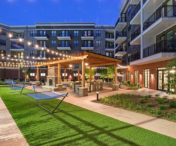 Enchanting apartment courtyard at dusk, featuring cozy seating under a wooden pergola, hanging string lights, modern landscaping, and inviting hammocks for relaxation.