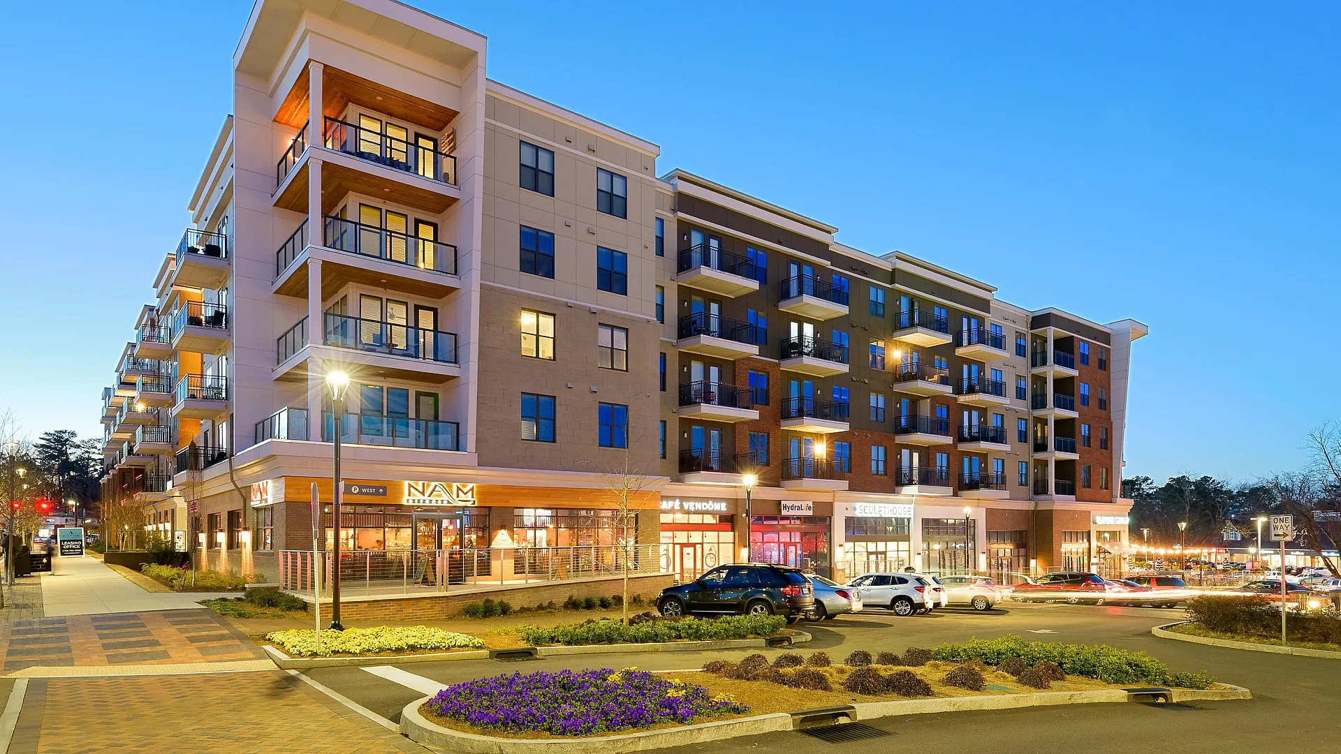 Modern apartment building with spacious balconies, vibrant street-level shops, and lively evening atmosphere.