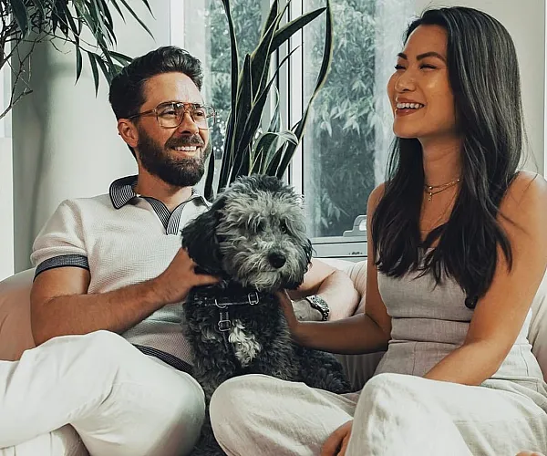 Happy couple relaxing with their fluffy dog on a cozy sofa in a bright, plant-filled living room.