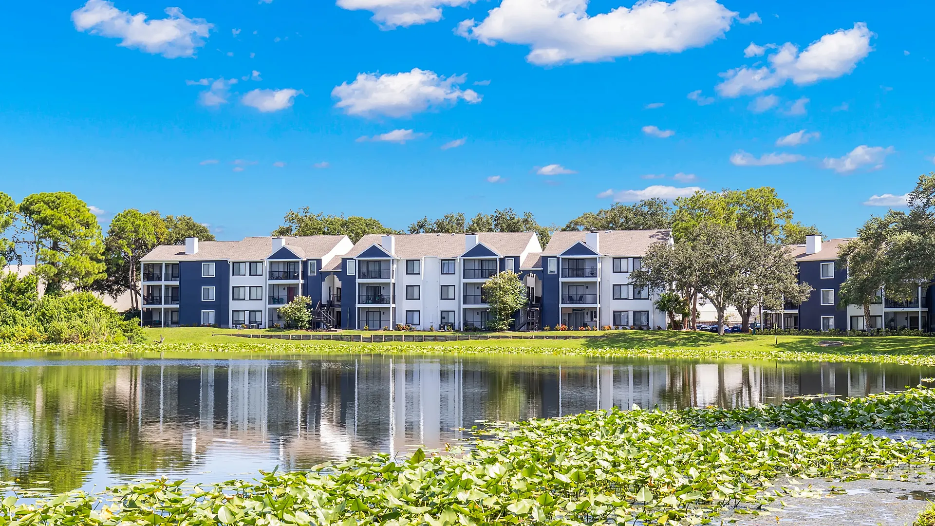 Charming lakeside apartment complex with modern architecture, set against a vibrant blue sky and lush greenery.