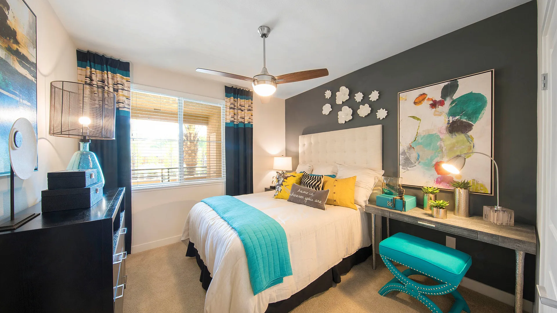 Bright and inviting bedroom with a chic design, featuring a plush bed adorned with vibrant pillows, a stylish ceiling fan, large window with natural light, and modern art decor.