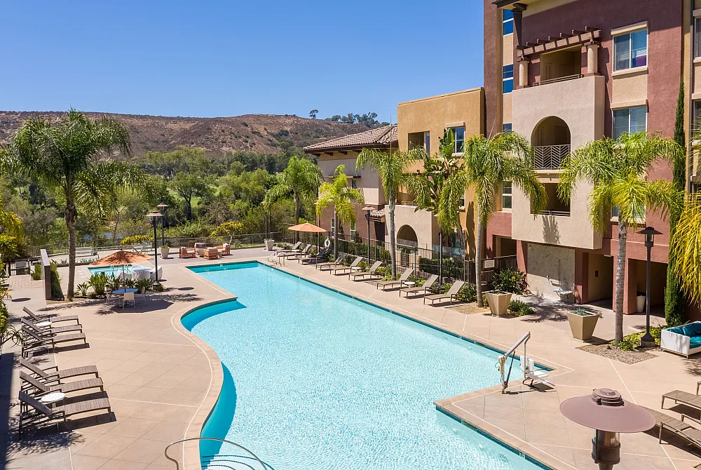 Luxurious apartment complex showcasing a sparkling outdoor pool, surrounded by lush palm trees and modern sun loungers, set against a scenic hillside background.