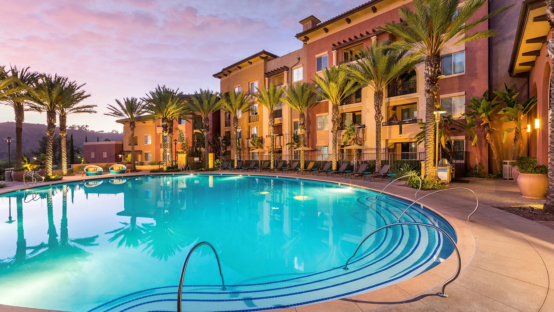 Luxury apartment complex with a stunning pool area, surrounded by palm trees and sunset skies, exuding a serene resort-like ambiance.