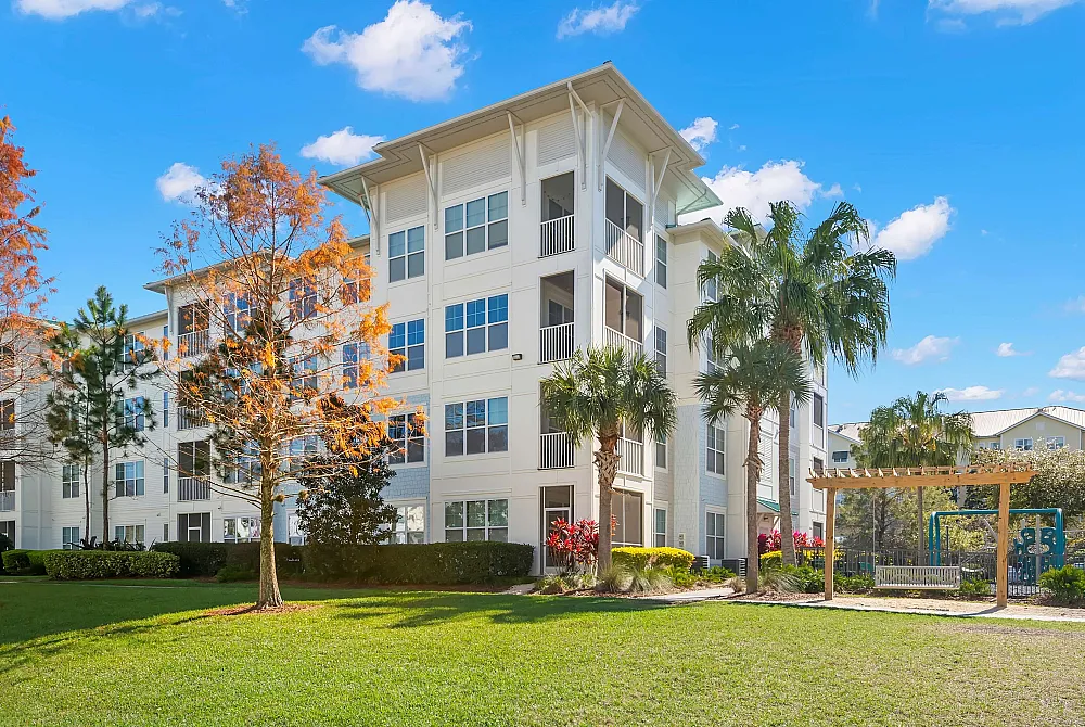 Charming apartment building surrounded by lush greenery and palm trees, featuring bright white exteriors and spacious balconies under a clear blue sky.