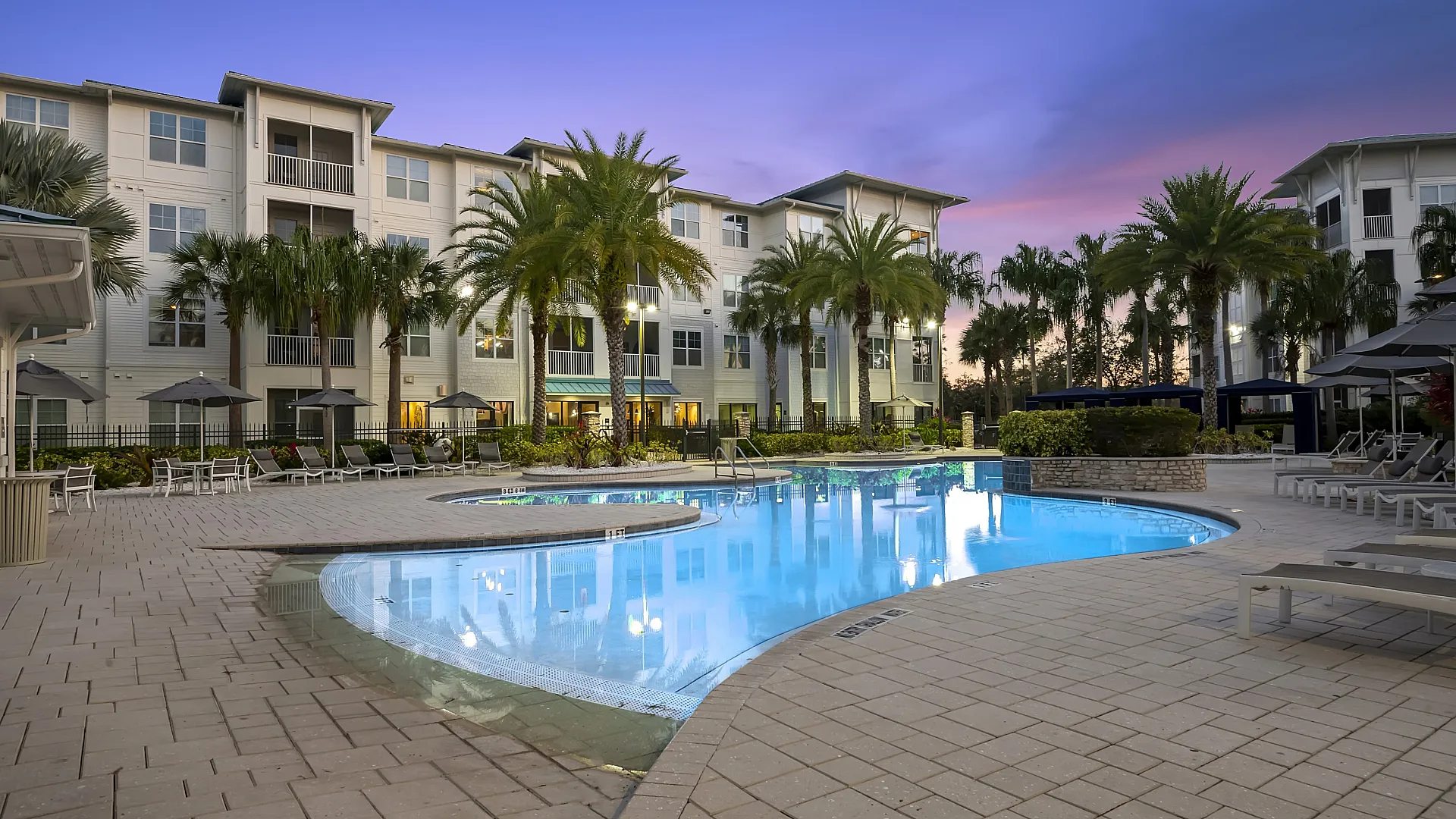 Luxurious apartment complex with modern architecture, featuring a pristine swimming pool surrounded by palm trees, creating a tropical oasis at sunset.