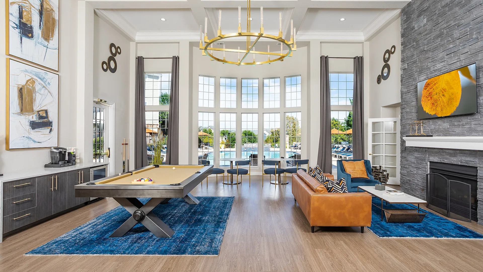 Luxurious and spacious living area with tall windows overlooking a pool, elegant chandelier, stylish pool table, and cozy seating arrangement on a chic blue rug.