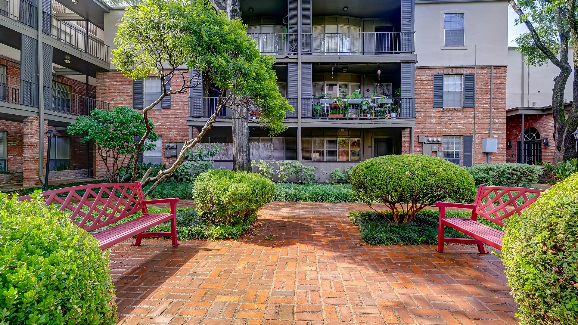 Charming apartment exterior with a brick facade, lush landscaping, and inviting red benches in a serene courtyard.