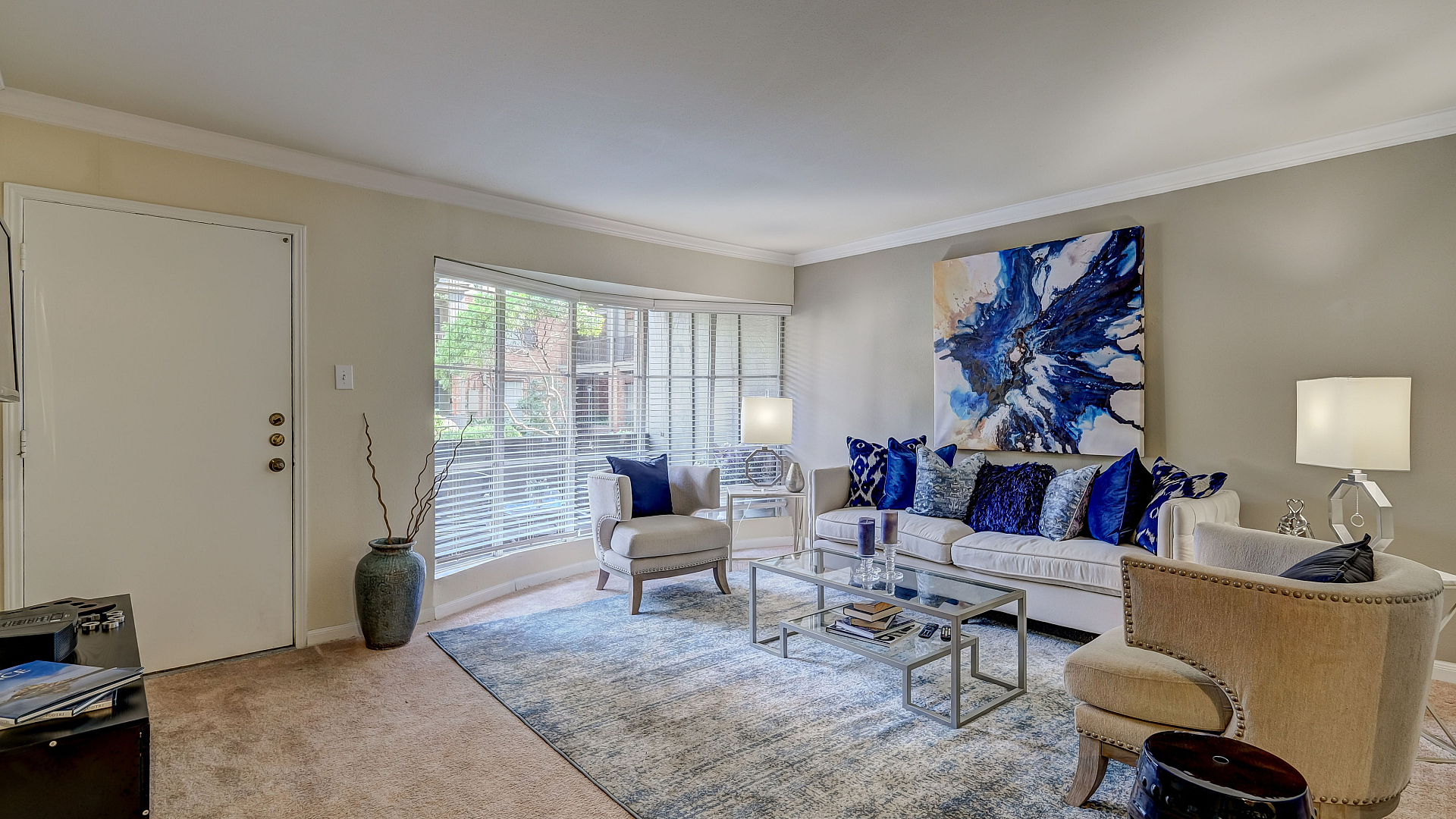 Elegant living room with chic decor, featuring a cozy white sofa, vibrant blue accents, modern glass coffee table, and natural light from a large window.