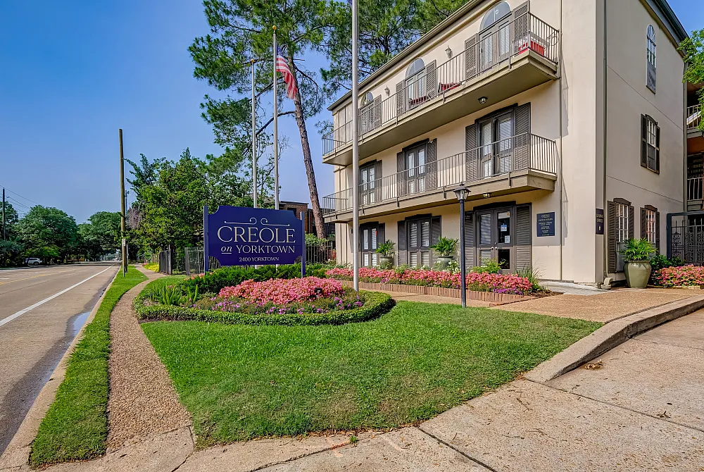 Charming Creole on Yorktown apartments showcasing elegant multi-level architecture with lush landscaping and vibrant flowers along a serene road.